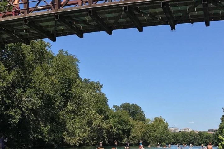 People on a bridge and in kayaks enjoy a sunny day on the calm river surrounded by trees.