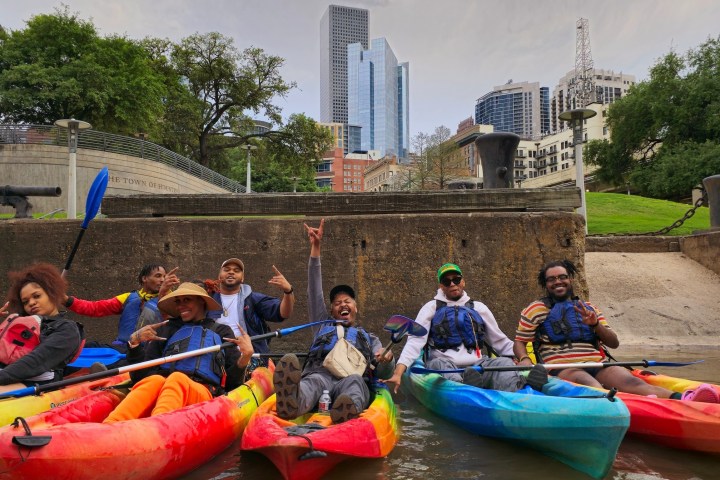 Group in colorful kayaks on a city waterway with buildings and trees in the background.