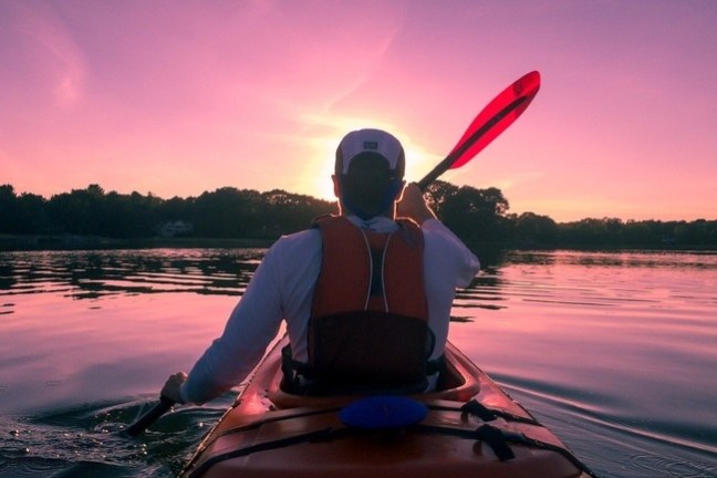 Person kayaking on a calm lake at sunset with a pink sky.