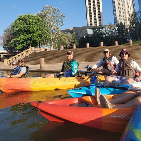 Group of people kayaking on a river in colorful kayaks with cityscape in the background.