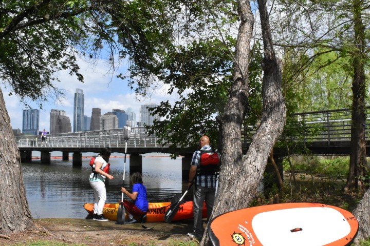 People near kayaks by a riverbank with city skyline and bridge in background.