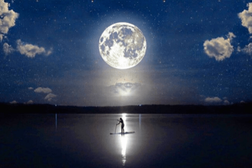 Person paddleboarding on a lake under a full moon at night with scattered clouds.