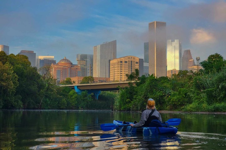 Person kayaking on river with city skyline in background under a cloudy sky.