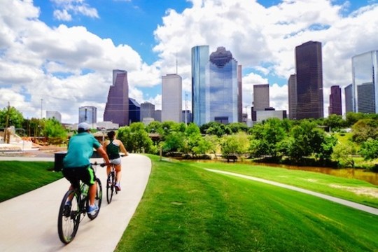 Two cyclists on a path with a city skyline and blue sky in the background.