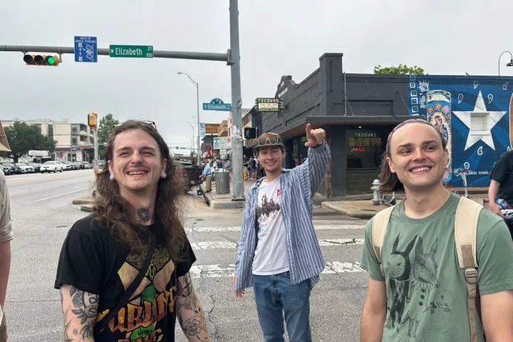 Three smiling people stand at a city intersection near a street sign.