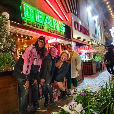 Four people smiling in front of neon-lit Dean's and Clarks signs on a lively city street at night.