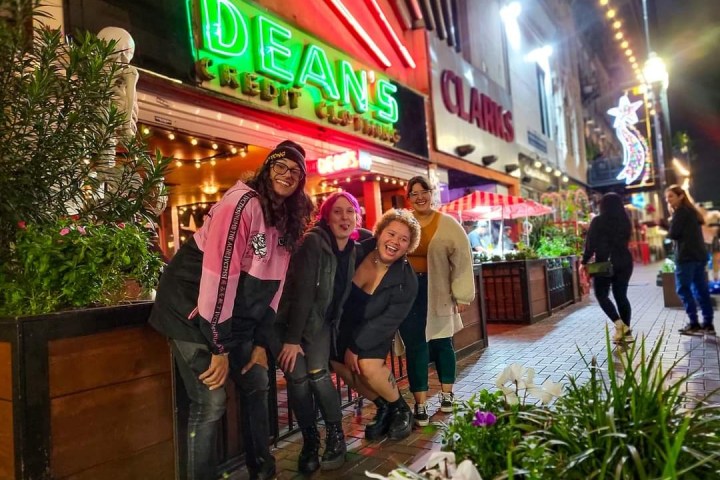 Four people smiling in front of neon-lit Dean's and Clarks signs on a lively city street at night.