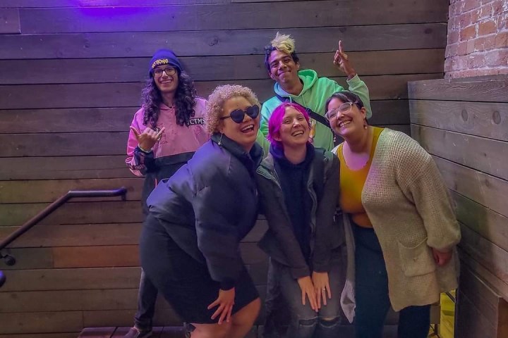 Five people posing under a bright neon arrow sign on a wooden staircase.