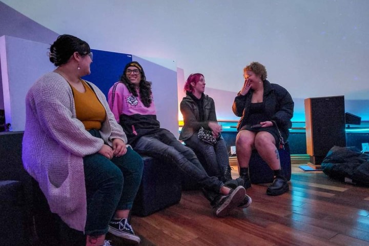 Four people sitting on chairs indoors, talking and laughing with soft lighting.