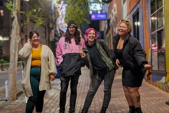 Four people smiling and posing on a brick street at night with colorful walls.