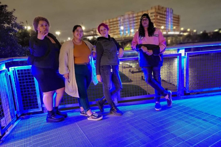 Four people stand on a brightly lit balcony with a city skyline at night.