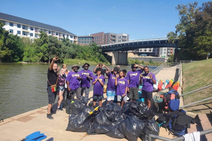 Group of people in purple shirts standing by river with trash bags and kayaks.