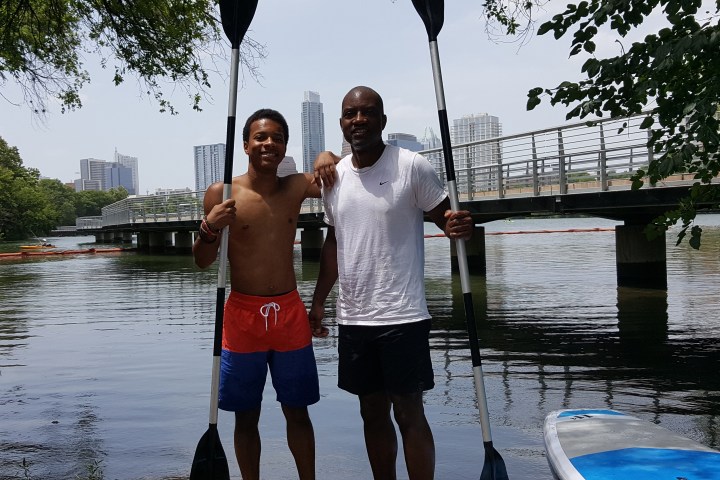 Two men holding paddles stand by a river with a bridge and city skyline in the background.