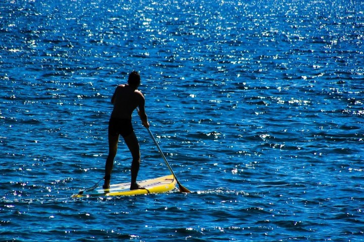 Person stand-up paddleboarding on calm, sunlit blue water.