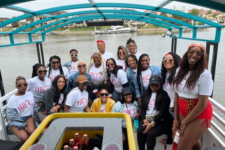 Group of people on a boat wearing matching HBCUs shirts, with water and houses in the background.