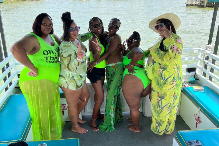 Six women posing on a boat, wearing bright green outfits, with water and docks in the background.