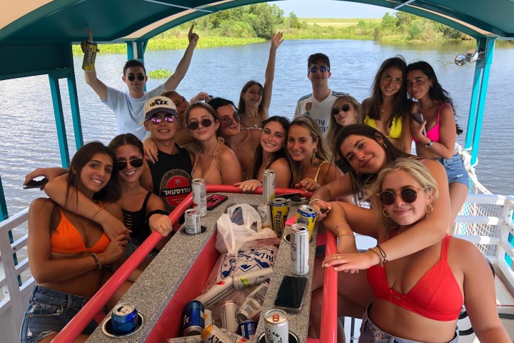 Group of friends on a boat, smiling and wearing swimwear, with drinks and snacks on a table.