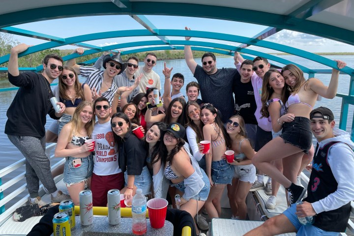 Group of young adults smiling and posing on a boat with drinks on a sunny day.