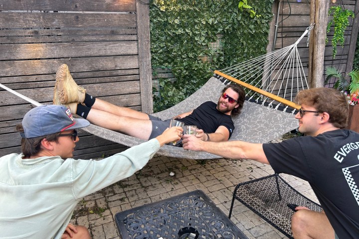 Three men toasting with drinks; one is lying on a hammock outdoors.