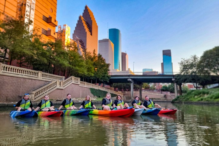 Group kayaking in urban river with city skyline and bridge in background.