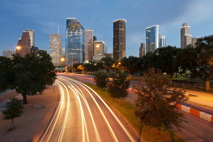 City skyline at dusk with light trails on the road and trees in the foreground.
