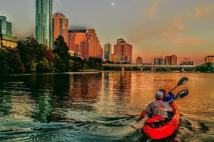 Person kayaking on a river with city skyline at sunset in the background.