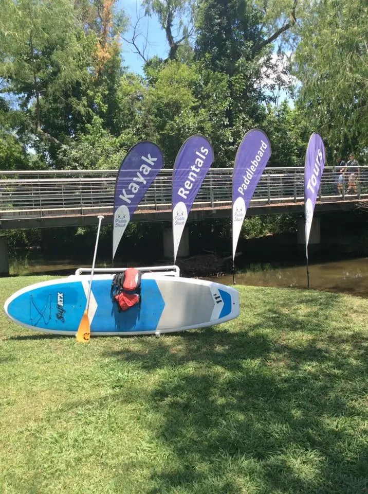 Paddleboard with paddle and gear on grass near river, flags read 'Kayak Rentals Paddleboard Tours'.