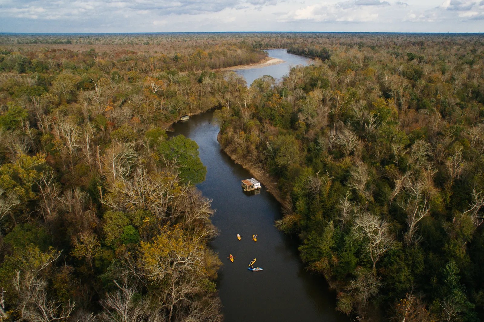 kayak-nola-wrenee-8 Aerial view of a river with kayaks and a houseboat surrounded by dense forest.